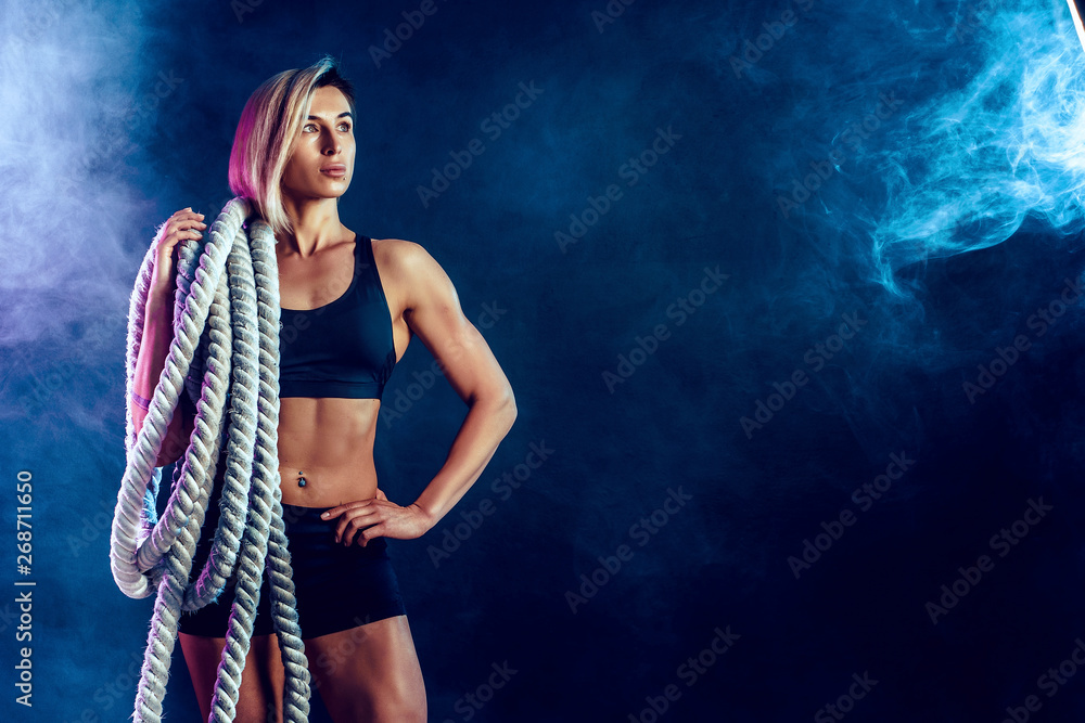 Attractive woman in black sportswear with heavy ropes on her shoulders on dark background. Strength and motivation. Sporty woman working with heavy ropes. Smoke on background.