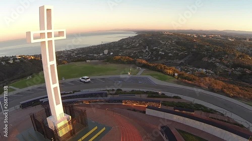 Reverse drone shot zooming past christian cross at a veterans memorial monument atop a mountain in urban setting overlooking the city and ocean with sweeping views over the horizon.