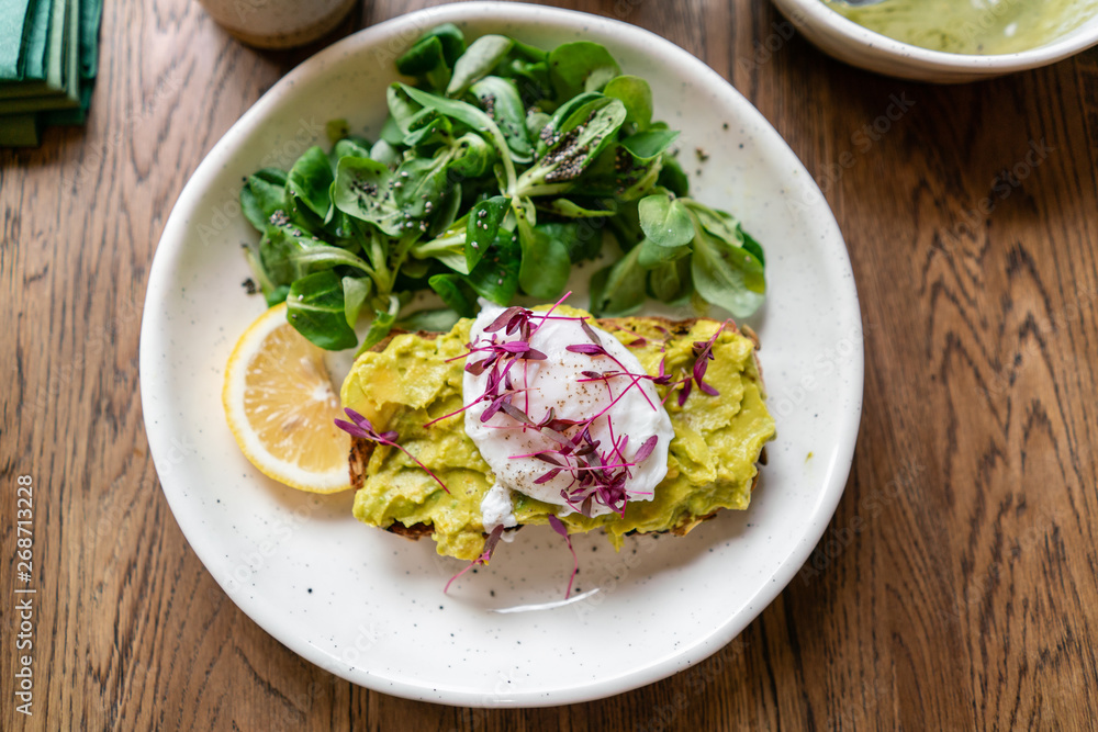 Morning in cafe, oak table. Healthy breakfast with wholemeal bread toast with avocado, poached egg with green salad. Green tea on the Background. Morning in cafe, oak table. Healthy breakfast with wholemeal bread toast with avocado, poached egg with green salad. Green tea on the Background.