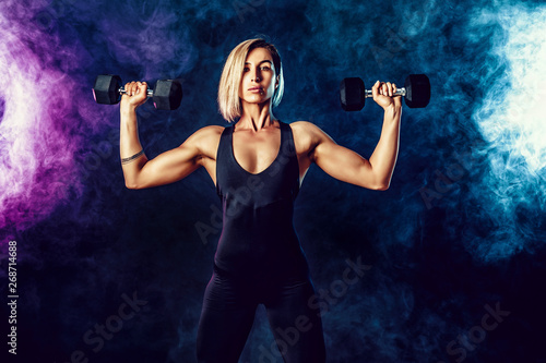 Wallpaper Mural Attractive sporty blonde woman in fashionable sportswear does the exercises with dumbbells. Photo of muscular woman on dark background with smoke. Strength and motivation. Torontodigital.ca
