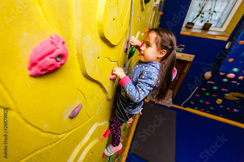 Little baby girl with funny hear style climbing vertical wall and man belaying her from below