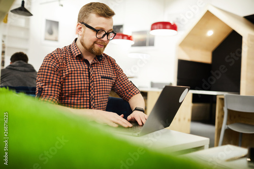 Content handsome chubby young man with beard sitting in coworking space and corresponding on laptop while working remotely