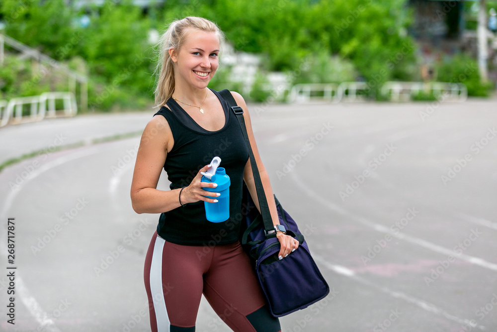 Fitness lifestyle. Young woman holding a water bottle and a bag on her shoulder after a workout. Workout at the stadium. Healthy life concept Fitness lifestyle. Young woman holding a water bottle and a bag on her shoulder after a workout. Workout at the stadium. Healthy life concept