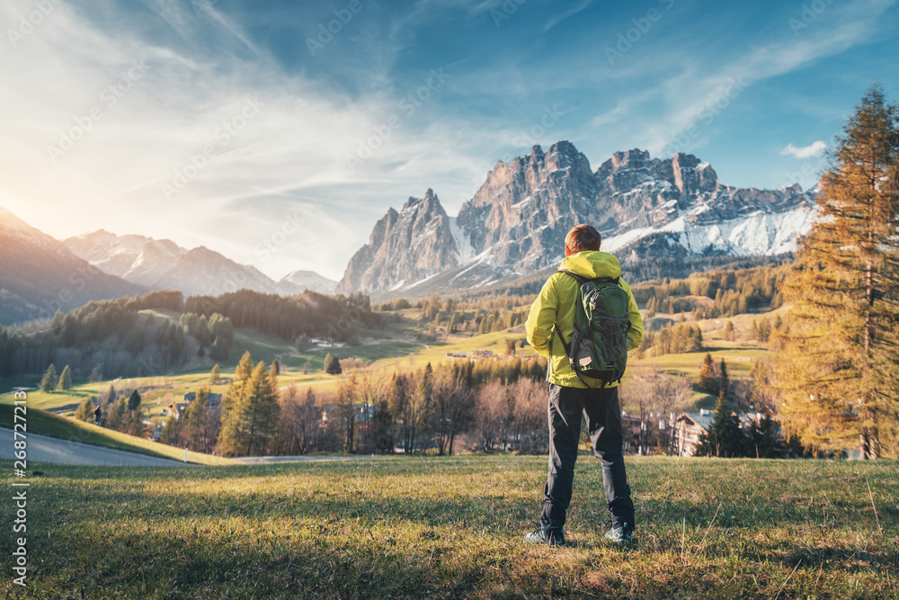 Young man in yellow jacket with backpack standing on the green meadow and looking on mountains at sunset in spring. Landscape with sporty guy, snowy rocks, trees, houses, blue sky in Italy. Travel