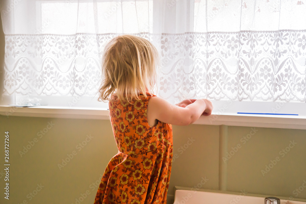 Young girl wearing orange dress with yellow flowers from upcycled old fabric. Alone exploring the bathroom of a 1970s house. Lace, sheer curtains and mustard yellow, green colored walls. Retro feel.
