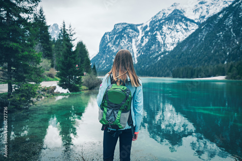 Young woman with green backpack is standing on the coast of mountain lake at cloudy day in spring. Travel in Dolomites, Italy. Landscape with girl, reflection in water, snowy rocks, trees. Vintage