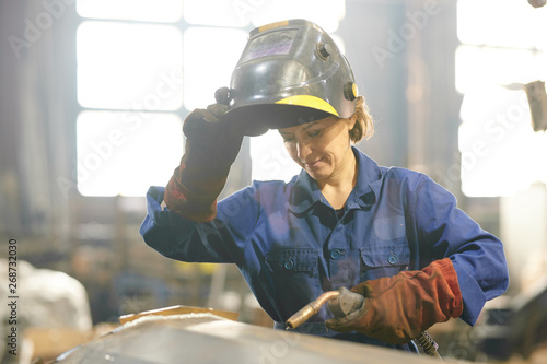 Tablou pe pânză Waist up portrait of smiling woman welding metal while working at industrial pla