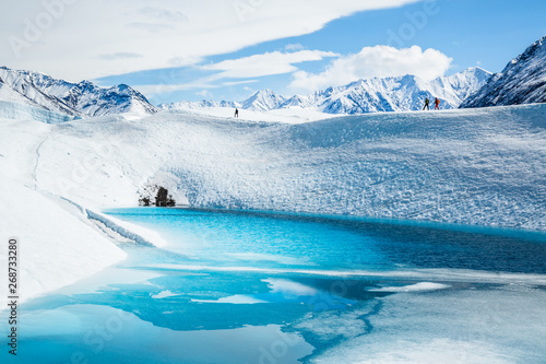 Three ice climbers trekking across a ridge of ice on the Matanuska Glacier in Alaska. They hike just above a steep slope over a deep blue pool.
