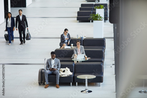 Serious tired business people in formal outfits sitting on couches and surfing net on modern gadgets while waiting for connecting flights in airport lounge