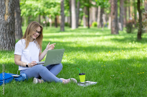Attractive young blonde woman-freelancer in glasses and earphones sit on the grass under tree in city park and listen music and look at laptop. Shows something with her hands and smiling