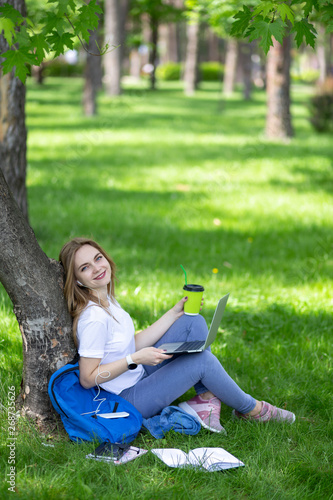 Charming young blonde woman in white t-shirt and jeans sit on the grass under tree in city park with notebook on her knees and cup of coffee in her hand. Listening music, looking at camera and smiling