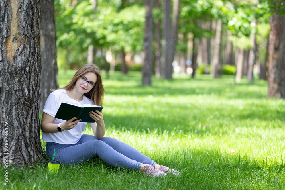 Joyful young blonde girl-student with glasses sits on grass under the tree and reads interesting book with smile. Green cup of coffee near. Student lifestyle. Space for text