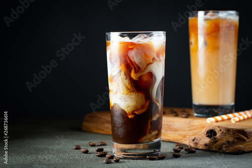 Fotografia Ice coffee in a tall glass with cream poured over, ice cubes and beans on a old rustic wooden table