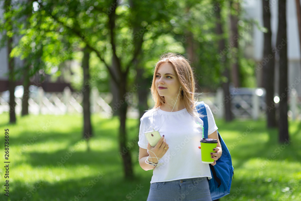 Woman in white t-shirt and blue jeans walks in the morning sunny park with dark-blue backpack and cup of coffee. She holds smartphone in her hand and listening music via earphones. Space for text Woman in white t-shirt and blue jeans walks in the morning sunny park with dark-blue backpack and cup of coffee. She holds smartphone in her hand and listening music via earphones. Space for text