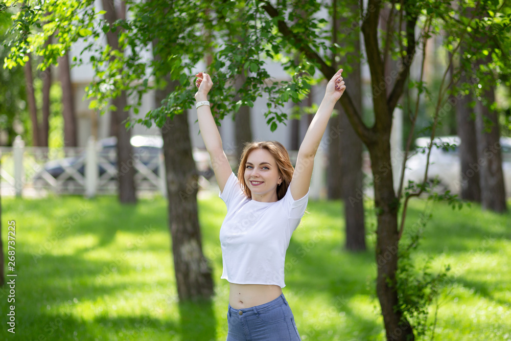 Happy smiling blonde girl in white t-shirt and blue jeans raised up her hands doing morning exercises in the sunny park.