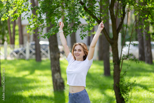 Happy smiling blonde girl in white t-shirt and blue jeans raised up her hands doing morning exercises in the sunny park.