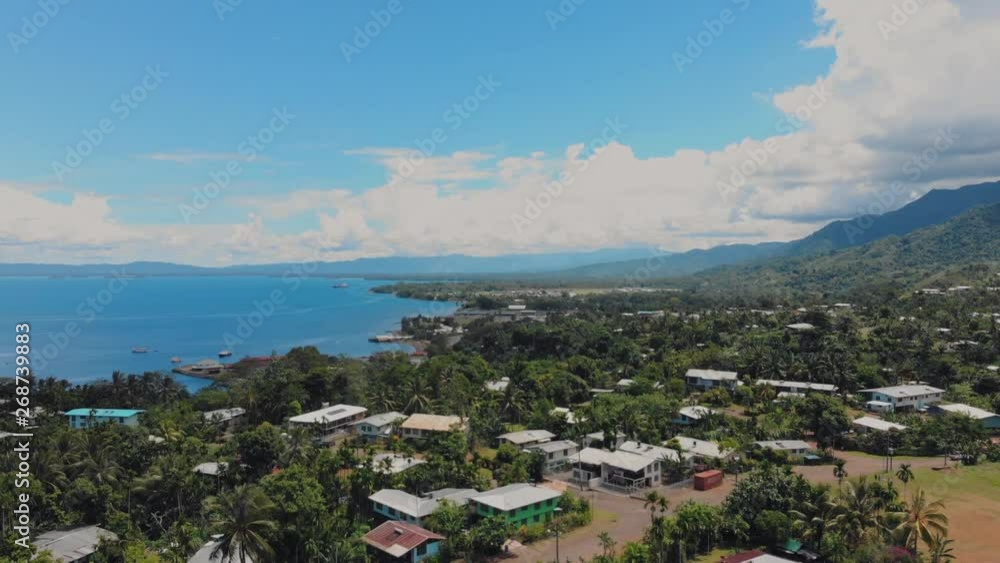 High, wide aerial view of Alotau, Papua New Guinea from a drone flying over the city toward the bright blue sea in the distance. Mountains rise up from the lush green landscape. High, wide aerial view of Alotau, Papua New Guinea from a drone flying over the city toward the bright blue sea in the distance. Mountains rise up from the lush green landscape.