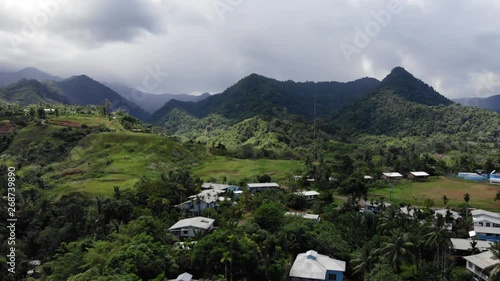 Drone ascending from the rugged tropical landscape of Alotau, Papua New Guinea, a tiny town on Milne Bay. Beautiful green terrain with houses tucked into the lush foliage with mountains and clouds. photo