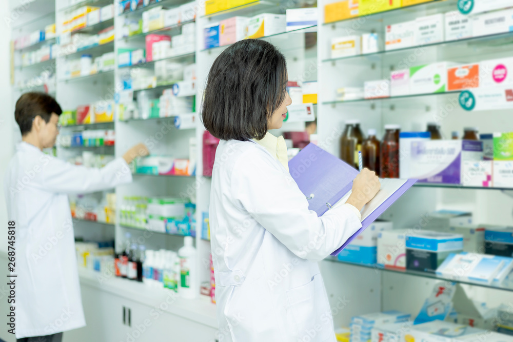 Junior Asian pharmacist medicine or checklist or writing on clipboard list of medicines and supplements with Senior Asian Woman pharmacist in drugstore or hospital pharmacy