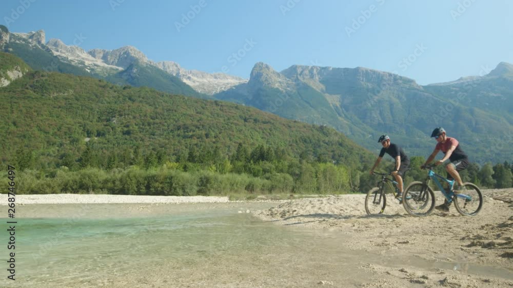 SLOW MOTION: Two fit guys ride their e-bikes up to a stream and observe the beautiful landscape. Young friends cross country cycling in Soca river valley look around the picturesque green mountains. SLOW MOTION: Two fit guys ride their e-bikes up to a stream and observe the beautiful landscape. Young friends cross country cycling in Soca river valley look around the picturesque green mountains.