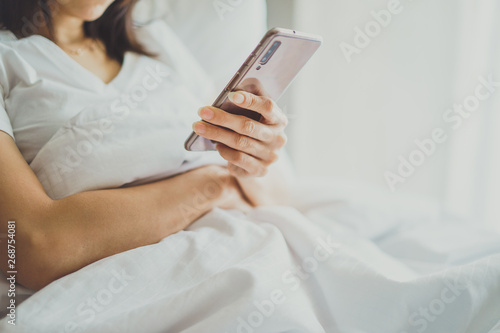 Asian Girl in bed room on the bed with the mobile phone.Closeup portrait of young sleepy exhausted woman lying in bed using smartphone 