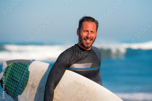 natural portrait of attractive and happy surfer man on his 40s carrying surf board after surfing morning at beautiful beach in wetsuit smiling enjoying Summer holiday