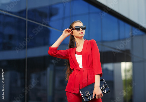 high fashion outdoor portrait of a young woman in a red pantsuit. Stylish black sunglasses, a black clutch bag, long hair.