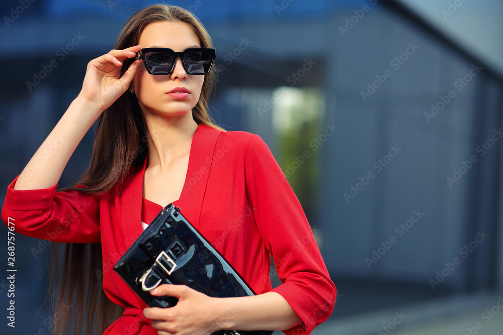 high fashion outdoor portrait of a young woman in a red pantsuit. Stylish black sunglasses, a black clutch bag, long hair. high fashion outdoor portrait of a young woman in a red pantsuit. Stylish black sunglasses, a black clutch bag, long hair.