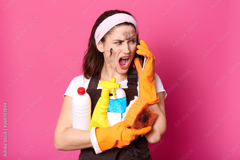 Image of screaming woman speaking on mobile phone while cleaning at home, dressed t shirt, apron and hairband, brunette lady holding sponge and detergents, model posing isolated over pink background. Image of screaming woman speaking on mobile phone while cleaning at home, dressed t shirt, apron and hairband, brunette lady holding sponge and detergents, model posing isolated over pink background.