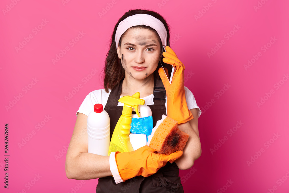 Close up shot of pretty housemaid wearing headband, white t shirt, brown and rubber protective gloves, holding cleaning materials, speaking via cell phone, posing isolated over pink background. Close up shot of pretty housemaid wearing headband, white t shirt, brown and rubber protective gloves, holding cleaning materials, speaking via cell phone, posing isolated over pink background.