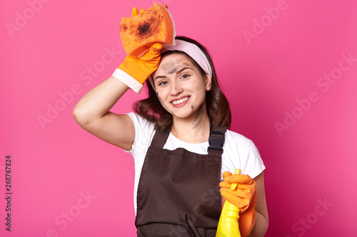Good looking housewife posing with hand touching head, holding detergent, smiling sincerely, having pleasant facial expression, posing isolated over pink background in studio. House chores concept. photo