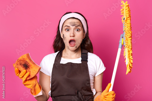Portrait of shocked exhausted cute woman with wide opened mouth and eyes, feeling of amazement, holding broom and dirty washcloth, being impressed, standing with dirty face. Household concept. photo