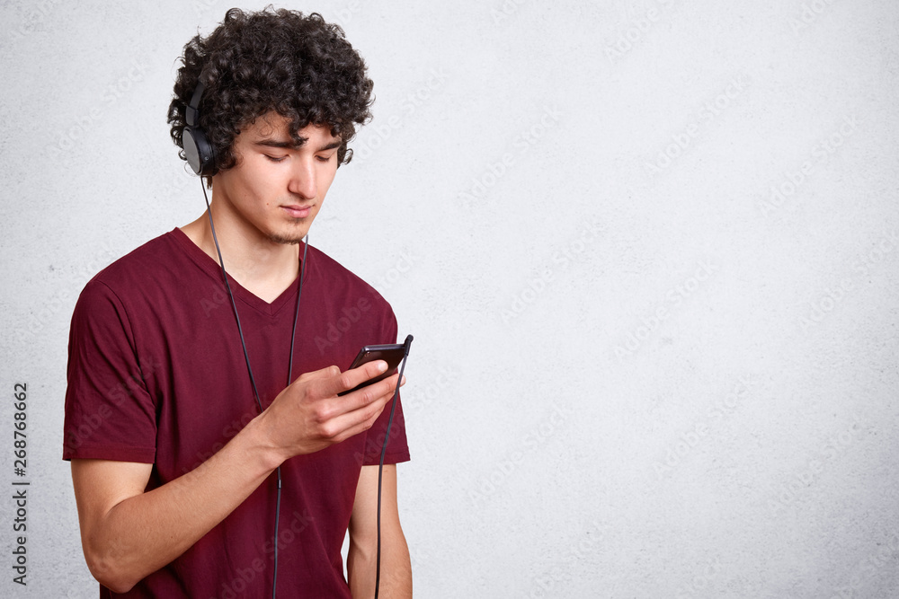 Horizontal shot of young handsome man, has dark curly hair, guy with cell  phone in hand