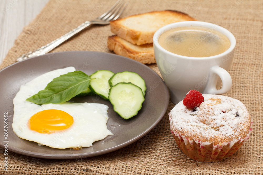 Plate with fried egg, fresh cut cucumber and leaf of basil, fork, cup of black coffee, cupcake and toasts. Plate with fried egg, fresh cut cucumber and leaf of basil, fork, cup of black coffee, cupcake and toasts.