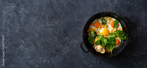Healthy breakfast table with fry pan eggs with spinach and corn on dark stone background top view