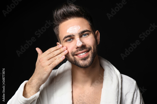 Handsome man applying clay mask on his face against dark background