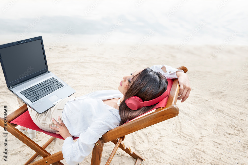Rear view of asian business woman relax when working with laptop while using headphones sitting in the beach chair on beach