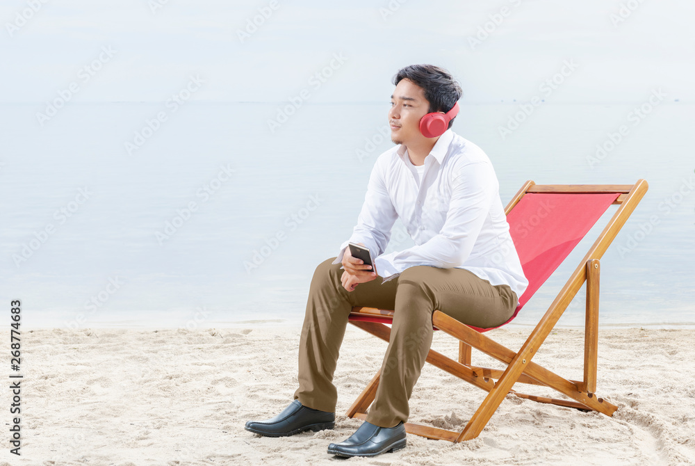 Asian businessman relax when working with mobile phone while using headphones sitting in the beach chair on beach
