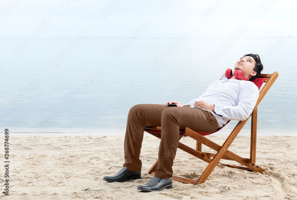 Asian businessman relax holding mobile phone while using headphones and lean back in the beach chair on beach