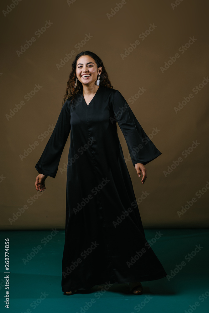Studio portrait of a beautiful and attractive Malay Asian woman in a traditional black baju kurung dress against a brown background. She is dancing and smiling as she poses for her photo. Studio portrait of a beautiful and attractive Malay Asian woman in a traditional black baju kurung dress against a brown background. She is dancing and smiling as she poses for her photo.