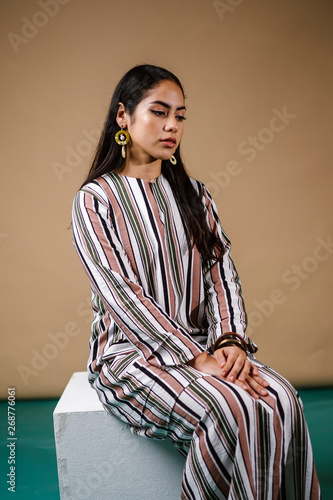 Studio portrait of a beautiful, tall, slim, elegant and young Middle Eastern woman in a traditional striped ethnic dress for Raya (Eid). She has a neutral expression as she poses for her headshot. 