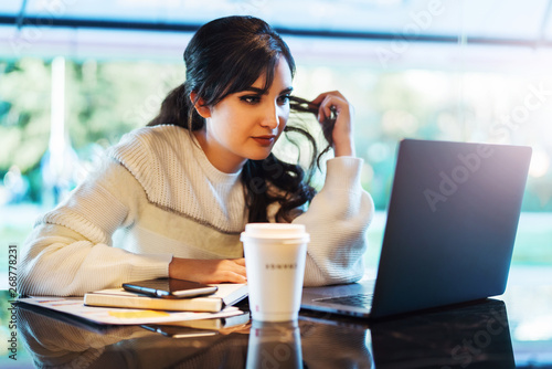 Portrait of young woman sitting at table in front of laptop. Girl works on computer cafe, checks e-mail, browses social networks.