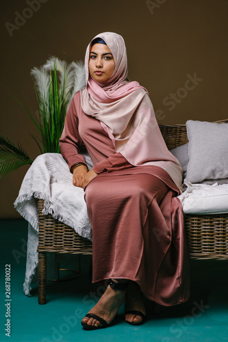 Studio portrait of a beautiful, vital and attractive young Middle Eastern Muslim woman in a pink baju kurung dress and hijab head scarf lounging on a sofa. She is dressed for visiting during Eid. photo
