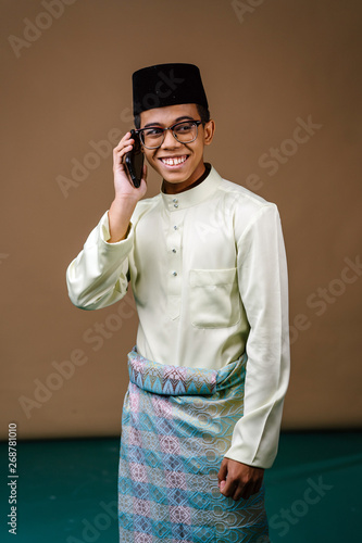 Studio portrait of a young and intelligent looking Malay Asian man in a baju melayu, sampin, songkok hat and spectacles. He is smiling as he uses his smartphone. He is dressed for Ramadan.  photo