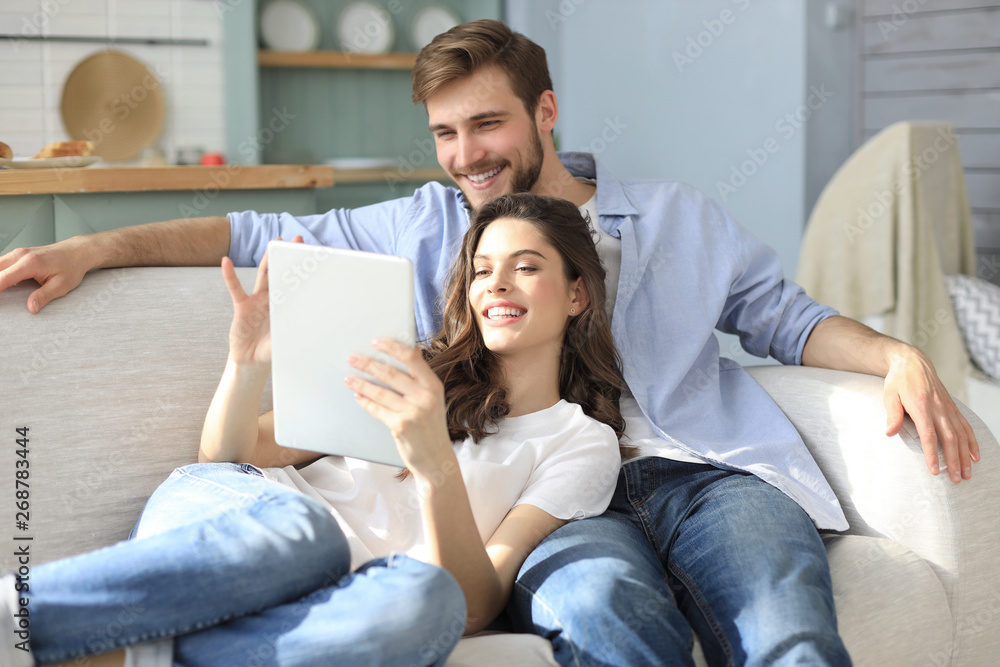 Young couple watching media content online in a tablet sitting on a sofa in the living room. - obrazy, fototapety, plakaty 