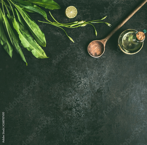 Wild garlic food background. Ramson leaves with bloom olives oil and wooden cooking spoon on dark rustic kitchen table, top view with copy space