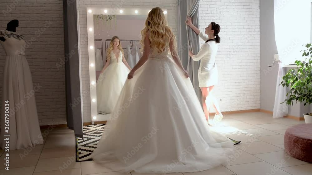 Wedding consultant helps bride choosen wedding dress in salon and wearing it near large mirror Wedding consultant helps bride choosen wedding dress in salon and wearing it near large mirror