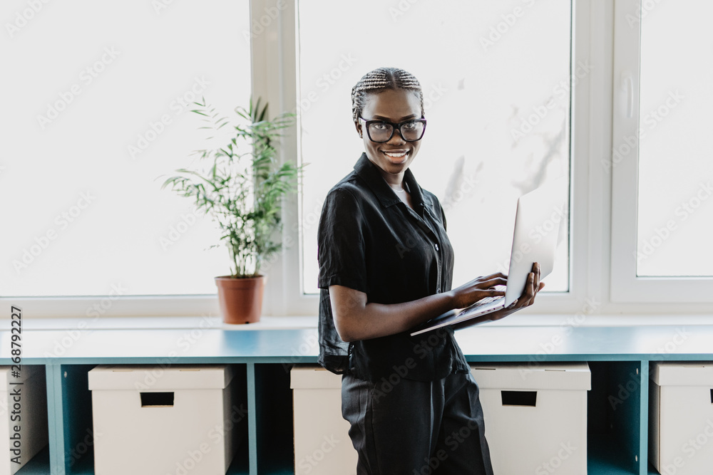Positive african woman in glasses standing and using laptop in modern office