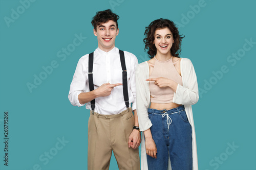 Portrait of happy successful two brunette stylish partner, couple or friends standing and pointing each other and looking at camera with toothy smile. indoor studio shot isolated on blue background.