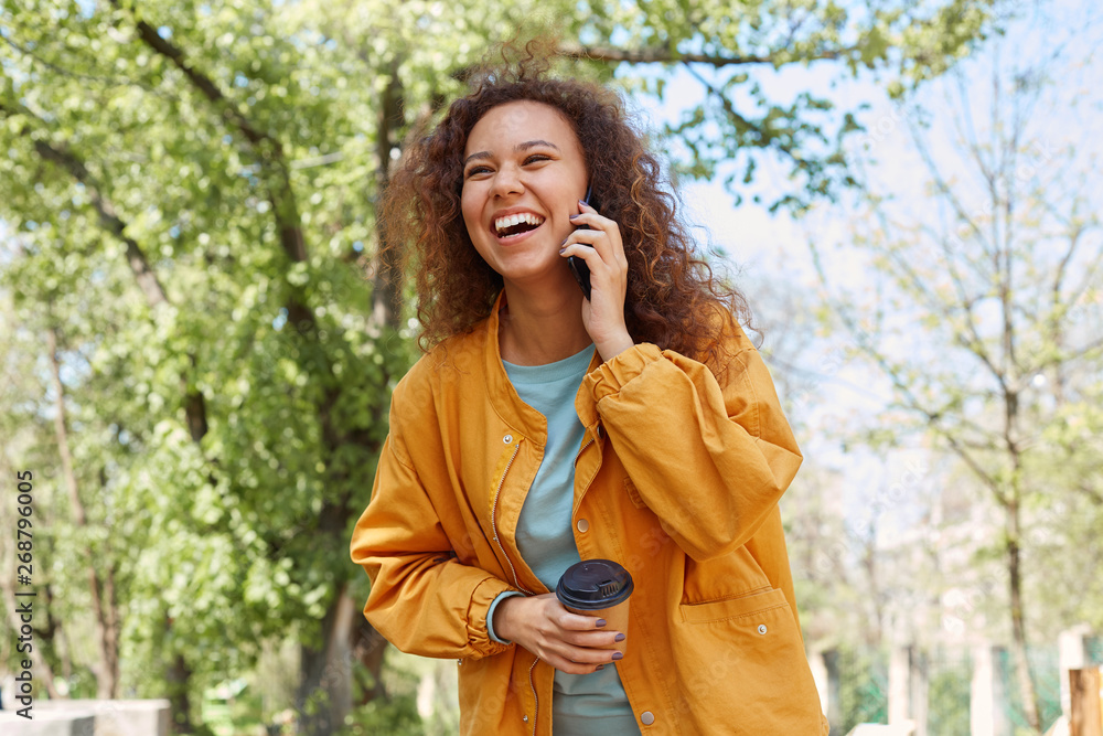 Happy laughing curly darck skined girl, wearing a yellow jacket, drinking coffee, enjoying the weather in the park, talking on the phone with his friend. Happy laughing curly darck skined girl, wearing a yellow jacket, drinking coffee, enjoying the weather in the park, talking on the phone with his friend.